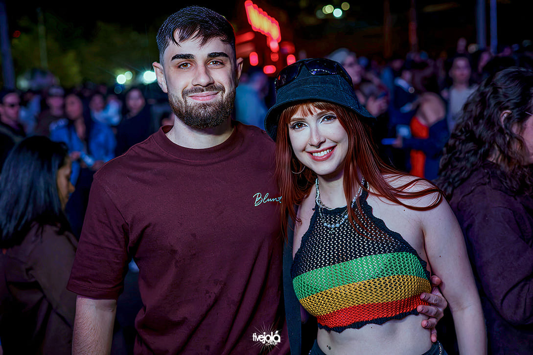 Festival Timbre - A imagem mostra um homem e uma mulher sorrindo e posando juntos. O homem, à esquerda, tem barba cheia, cabelo curto e escuro, e veste uma camiseta marrom com a palavra "Blunt" bordada no peito. A mulher, à direita, tem cabelo ruivo liso, usa um chapéu bucket preto e óculos escuros apoiados sobre ele. Ela veste um top de crochê preto com listras verde, amarela e vermelha, além de um colar prateado.